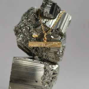 Gold necklace displayed on shiny geometric mineral, architectural t-bar pendant made from gold, with hand-carved texture resembling mineral stritations