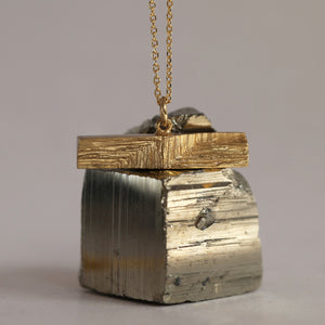 Bocky gold pendant leaning on shiny mineral, geometric gold t-bar pendant suspended on a gold chain, decorated with etched texture resembling mineral stritations