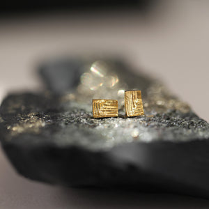 Small gold earrings on shiny rock surface, rectangular dainty gold stud earrings with intricate texture resembling mineral stritations