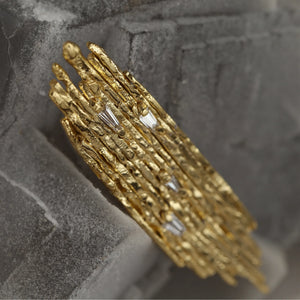 Amorphe modernist-inspired statement brooch in fire-textured 18kt gold and white tapered baguette diamonds on a grey mineral, angled view.